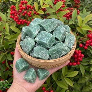 May include: A hand holds a woven basket filled with rough-cut green aventurine stones. Several stones are also held in the hand. The background features green leaves and red berries.