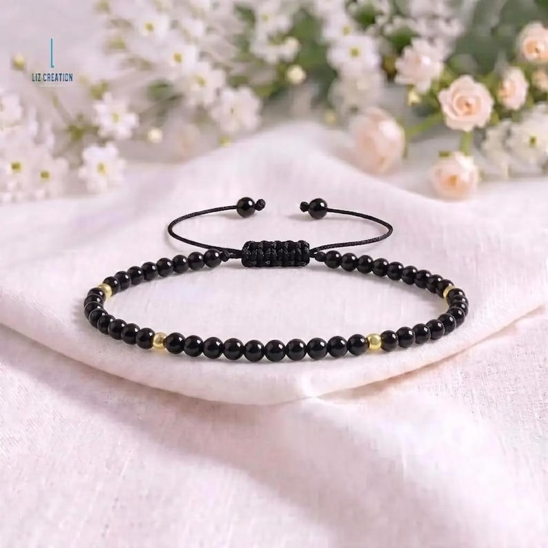 May include: A black beaded bracelet featuring small, round, polished black beads interspersed with two metallic gold-colored beads. The bracelet has an adjustable black cord closure with a woven knot mechanism. The text LIZ CREATION is visible.