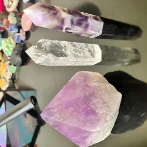 Puede incluir: Tres puntas de cristal de amatista, una con base negra, una punta de cuarzo transparente y un gran cristal de amatista. Los cristales est&aacute;n sobre una superficie blanca.