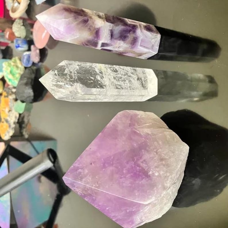 Puede incluir: Tres puntas de cristal de amatista, una con base negra, una punta de cuarzo transparente y un gran cristal de amatista. Los cristales est&aacute;n sobre una superficie blanca.