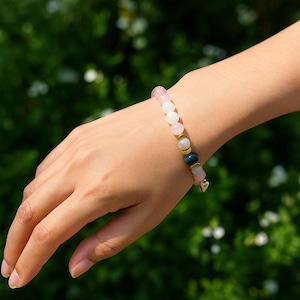 Pode incluir: Uma pulseira de contas rosa, brancas e verde-azuladas, acentuada com contas douradas e um pequeno pingente de cora&ccedil;&atilde;o. A pulseira &eacute; usada no pulso contra um fundo verde desfocado.