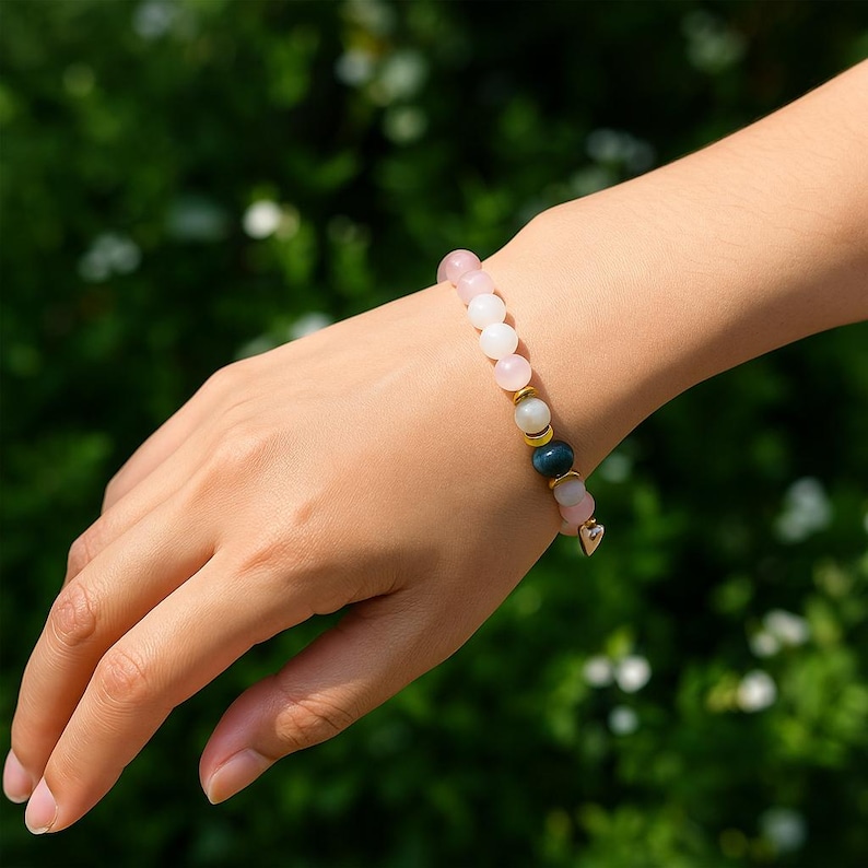 Pode incluir: Uma pulseira de contas rosa, brancas e verde-azuladas, acentuada com contas douradas e um pequeno pingente de cora&ccedil;&atilde;o. A pulseira &eacute; usada no pulso contra um fundo verde desfocado.