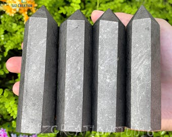 Può includere: Quattro obelischi di cristallo sfaccettati grigio scuro sono tenuti in una mano. Ogni obelisco ha una sommità appuntita e un corpo rettangolare. I cristalli sono di un colore grigio tenue e sembrano essere fatti di pietra. Lo sfondo è un fogliame verde sfocato.
