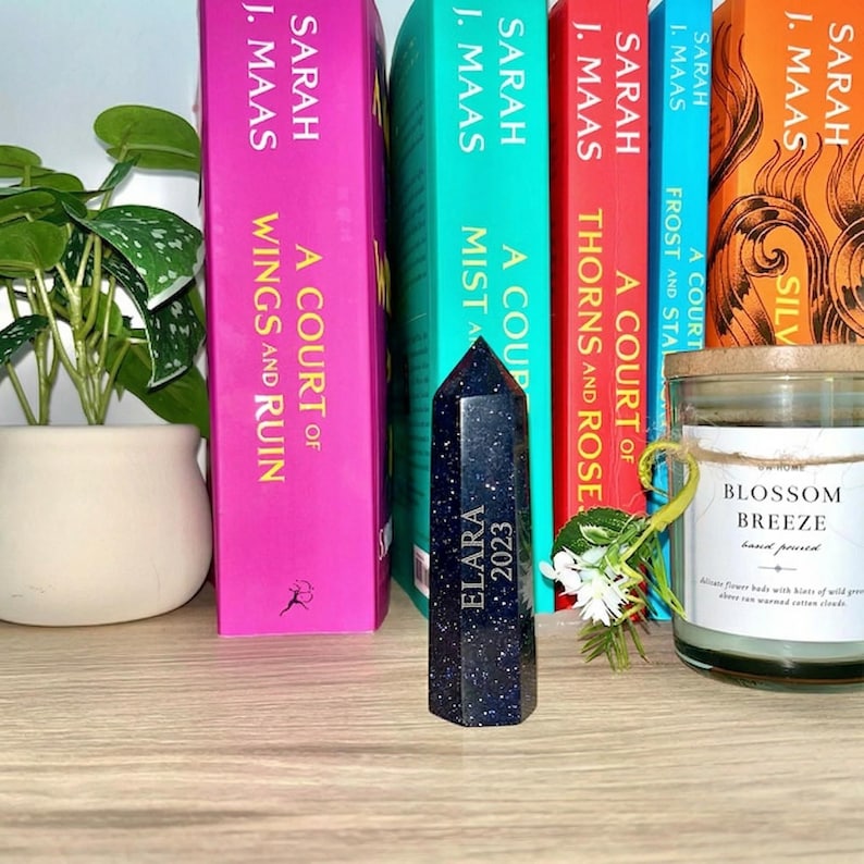 May include: A dark blue crystal obelisk with the text "ELARA 2023" stands in front of a row of books. The books have colorful spines and the author's name "SARAH J. MAAS" is visible. A candle and a potted plant are also in the image.