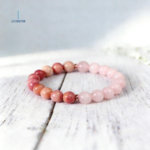 Puede incluir: Un brazalete de cuentas con piedras preciosas de tonos rosas y rojos. El brazalete presenta un degradado de color, pasando de cuentas rosa-rojizas oscuras a cuentas rosa pastel m&aacute;s claras. El texto "LIZ CREATION" es visible.