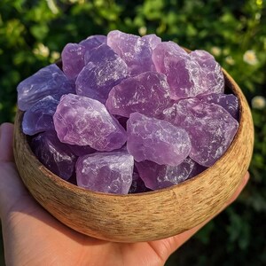 May include: A wooden bowl filled with raw amethyst crystals. The crystals vary in shades of purple, from light lavender to deep violet. The bowl is made of light-colored wood, and the background is blurred green foliage.