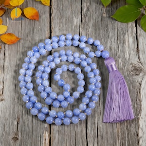 Pode incluir: Um colar mala de ágata azul rendada com borla lavanda. O colar é feito de contas redondas azuis claras com marmoreio branco. A borla é de cor lavanda lisa. O colar é exibido em uma superfície de madeira envelhecida.