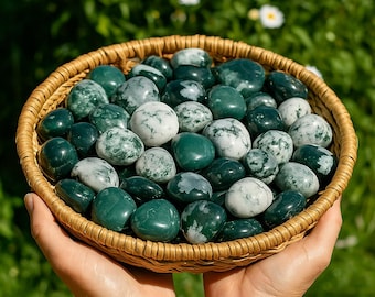 May include: A woven basket filled with polished green and white moss agate stones. The stones vary in size and have a speckled appearance. The basket is held in two hands against a blurred green background.
