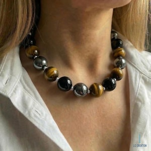 May include: A beaded necklace featuring alternating polished beads in black, silver, and tiger's eye. The necklace is strung on a white cord and is worn against a white shirt.