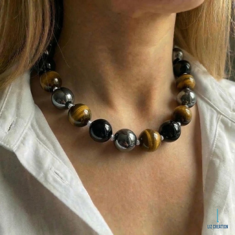 May include: A beaded necklace featuring alternating polished beads in black, silver, and tiger's eye. The necklace is strung on a white cord and is worn against a white shirt.