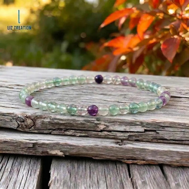 May include: A beaded bracelet featuring translucent green, purple, and clear beads. The bracelet is strung on a clear elastic cord and rests on a weathered wooden surface. The text "LIZ CREATION" is visible in the upper left corner.