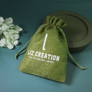 Op de afbeelding: Een groene jute zak met trekkoord en witte tekst met de tekst "LIZ CREATION" en "HEAL THE MIND, BODY AND SOUL". De zak is aan de bovenkant vastgebonden met een bijpassend groen trekkoord. De achtergrond is donkergroen.