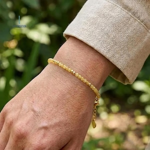 Op de afbeelding: Een delicate gouden armband met kleine, ronde, gele kralen. De armband zit om een pols, met een gouden sluiting en een kleine bedel. De achtergrond is wazig, met groen gebladerte en een beige mouw.