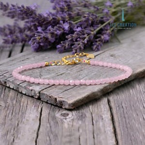 May include: A delicate pink beaded bracelet with a gold-colored clasp and accents. The bracelet is displayed on a weathered wooden surface, with a backdrop of purple lavender sprigs. The bracelet's beads are small and faceted, reflecting light.