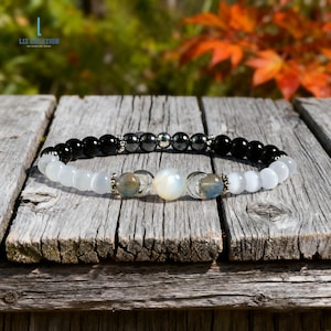 Puede incluir: Una pulsera de cuentas negras, blancas y plateadas, con una sección central de piedras iridiscentes y azul-gris. La pulsera se exhibe sobre una superficie de madera envejecida, con un fondo borroso de follaje otoñal.