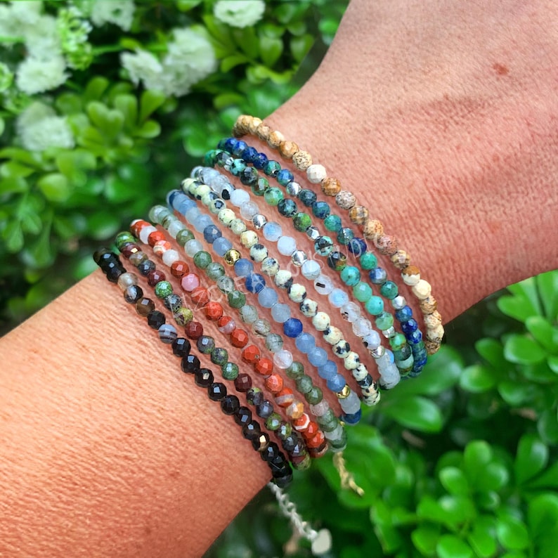 May include: A stack of ten thin bracelets made of small, faceted, multi-colored gemstone beads. The bracelets feature various colors including black, dark green, red, light green, blue, white, turquoise, and tan, arranged in a layered design.