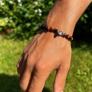 Pode incluir: Uma pulseira de contas com contas marrons, pretas e douradas. Possui um pequeno pingente escuro e uma conta azul clara. A pulseira &eacute; usada no pulso.