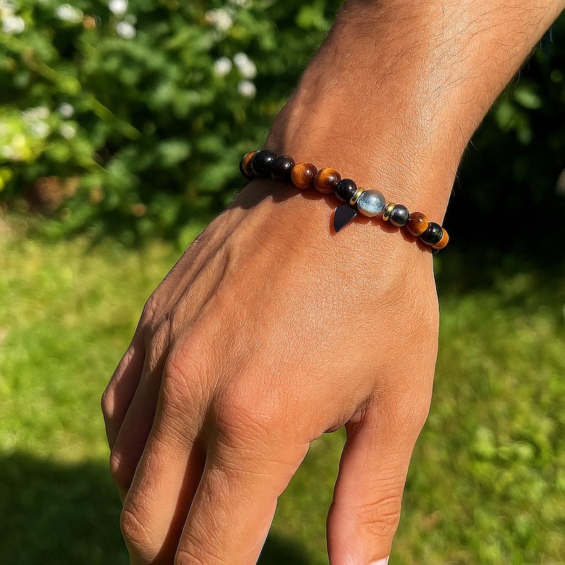 Pode incluir: Uma pulseira de contas com contas marrons, pretas e douradas. Possui um pequeno pingente escuro e uma conta azul clara. A pulseira &eacute; usada no pulso.