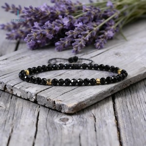 May include: Black beaded bracelet with gold-colored accents. The bracelet is made of small, faceted black beads and features two gold-colored square beads. The bracelet is displayed on a weathered wooden surface, with a sprig of purple lavender in the background.