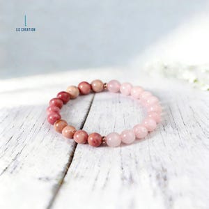 Puede incluir: Una pulsera de cuentas con piedras preciosas rosas y rojas, dispuestas en degradado. Las cuentas pasan del rojo oscuro al rosa p&aacute;lido. La pulsera est&aacute; sobre una superficie de madera blanca. El texto "LIZ CREATION" es visible en la esquina superior izquierda.