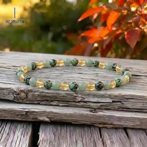 May include: A beaded bracelet featuring a mix of green, yellow, and speckled beads. The bracelet is displayed on a weathered wooden surface, with the text "LIZ CREATION" visible in the upper left corner. The background includes blurred greenery and red foliage.