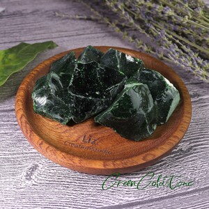 Puede incluir: Un cuenco de madera lleno de varios trozos de vidrio verde oscuro. Los trozos de vidrio tienen bordes &aacute;speros e irregulares. El cuenco es de color marr&oacute;n c&aacute;lido y est&aacute; sobre una superficie gris claro. El texto "Liz Creation Studio" es visible.