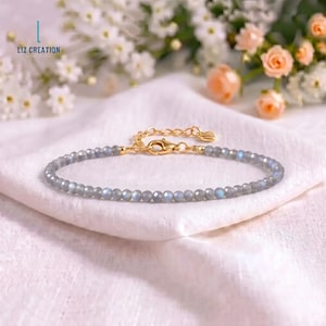 May include: A delicate bracelet featuring small, faceted grey labradorite beads with iridescent blue flashes. The bracelet includes a gold-toned metal chain, a lobster claw clasp, and a small heart-shaped charm at the end of the extender chain.