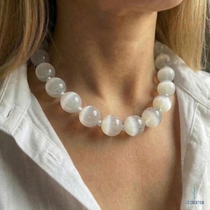 May include: A white selenite beaded necklace. The necklace features large, round, polished beads strung together. The beads have a translucent, milky appearance. The necklace is worn against a white shirt.