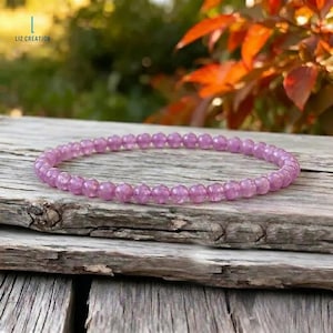 Op de afbeelding: Een delicate, lichtpaarse kralenarmband ligt op een verweerd houten oppervlak. De armband bestaat uit kleine, ronde kralen, met een consistente kleur en textuur. De achtergrond toont wazig groen en herfstbladeren, wat een natuurlijke esthetiek toevoegt.