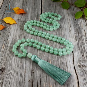 May include: Three strands of green gemstone beads with a matching green tassel. The beads are round and smooth, and the tassel is made of silky threads. The beads are displayed on a weathered wooden surface.