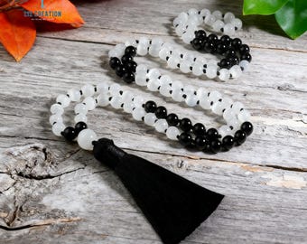 Può includere: Una collana di perline con perline bianche e nere alternate, con una grande perla bianca e un nappa nera. La collana è esposta su una superficie di legno invecchiato, con accenni di fogliame verde e arancione sullo sfondo.