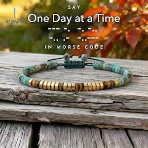 May include: A beaded bracelet with turquoise, gold, and brown beads. The bracelet is on a wooden surface. The text on the image says "One Day at a Time" in Morse code.