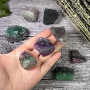 Puede incluir: Una colecci&oacute;n de cristales de fluorita en bruto en tonos morados, verdes y blancos. Los cristales se exhiben sobre una superficie de madera, algunos sostenidos en una mano. Se ven ramitas de lavanda seca en el fondo.