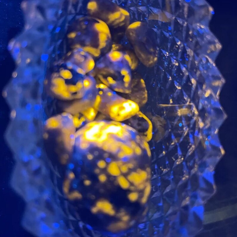 May include: A collection of tumbled stones with a dark blue and yellow pattern, filling a clear, faceted glass container. The stones have a speckled appearance, and the container has a diamond-like design.
