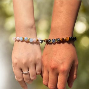 Pode incluir: Pulseiras de contas combinando com um pingente de cora&ccedil;&atilde;o magn&eacute;tico. Uma pulseira apresenta contas rosa e azuis com detalhes dourados, enquanto a outra tem contas marrons, pretas e douradas. As pulseiras est&atilde;o conectadas, simbolizando um v&iacute;nculo.