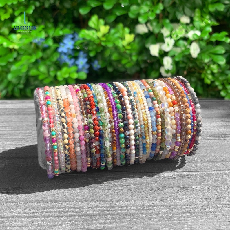 May include: A collection of numerous thin beaded bracelets arranged in a row, featuring a wide variety of colorful gemstones and small metallic accents. Each bracelet consists of small, round, polished beads in shades of pink, blue, green, yellow, and clear, creating a vibrant spectrum of colors displayed on a gray surface.