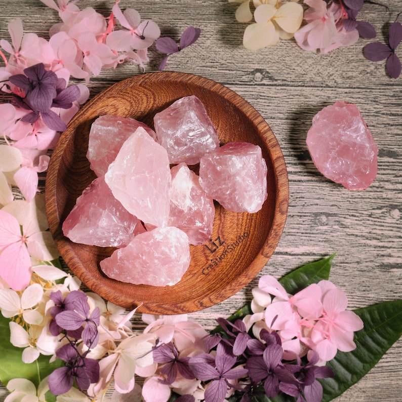 May include: A wooden bowl filled with raw, light pink rose quartz crystals. The crystals vary in size and shape, with a rough, natural texture. The bowl is surrounded by pink and purple dried flowers, creating a soft, natural aesthetic.