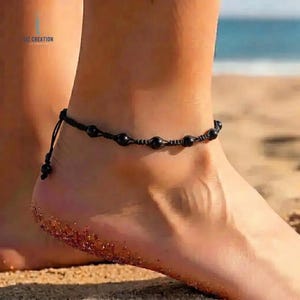 May include: Black beaded anklet with a macrame design. The anklet features several black beads and is worn on an ankle. The background shows a beach setting with sand and water.