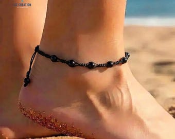 May include: Black beaded anklet with a macrame design. The anklet features several black beads and is worn on an ankle. The background shows a beach setting with sand and water.