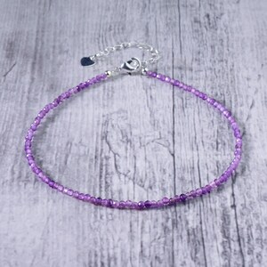 May include: A delicate purple beaded bracelet with a silver clasp and extender chain. The bracelet is made of small, round beads and is displayed on a light gray wooden surface. The clasp has a small heart charm.