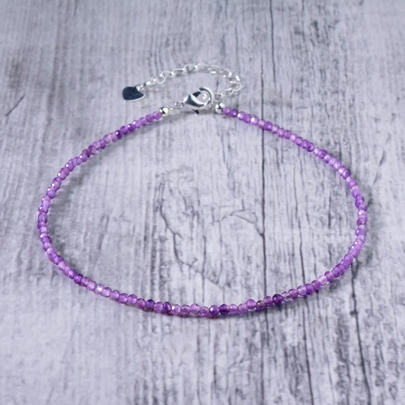 May include: A delicate purple beaded bracelet with a silver clasp and extender chain. The bracelet is made of small, round beads and is displayed on a light gray wooden surface. The clasp has a small heart charm.