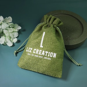 May include: A green burlap drawstring bag with white text reading "LIZ CREATION" and "HEAL THE MIND, BODY AND SOUL". The bag is on a dark green surface with a matching decorative element and white flowers.