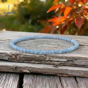 May include: A delicate, light blue beaded bracelet rests on a weathered wooden surface. The bracelet is composed of small, round beads, creating a subtle, elegant accessory. The text "LIZ CREATION" is visible in the upper left corner.