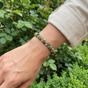 May include: A braided bracelet featuring green, brown, and gold beads. The bracelet is worn on a wrist, with a neutral-colored sleeve in the background. The bracelet has a drawstring closure.