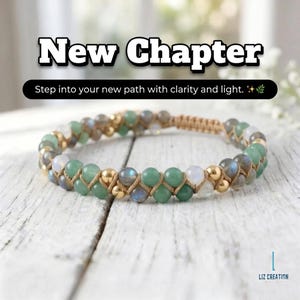 May include: A beaded bracelet with green, gray, and gold beads, woven with a tan cord. The bracelet is on a white wooden surface. Text above the bracelet reads "New Chapter" and "Step into your new path with clarity and light."