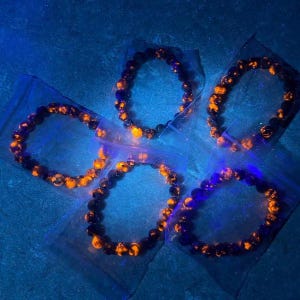 Puede incluir: Cinco pulseras hechas de cuentas oscuras con acentos naranjas se muestran en bolsas de pl&aacute;stico transparente individuales. Las pulseras est&aacute;n dispuestas sobre una superficie azul, y la iluminaci&oacute;n da a la imagen un efecto brillante.