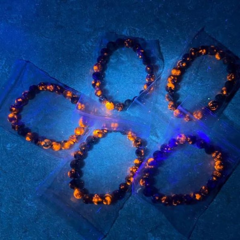 Puede incluir: Cinco pulseras hechas de cuentas oscuras con acentos naranjas se muestran en bolsas de pl&aacute;stico transparente individuales. Las pulseras est&aacute;n dispuestas sobre una superficie azul, y la iluminaci&oacute;n da a la imagen un efecto brillante.