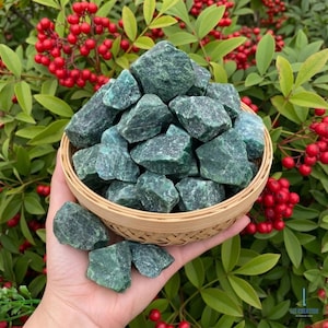 May include: A collection of rough, green gemstones in a small, woven basket. The stones are a deep, forest green color and have a textured surface. The basket is held in a hand, with a backdrop of green leaves and red berries.