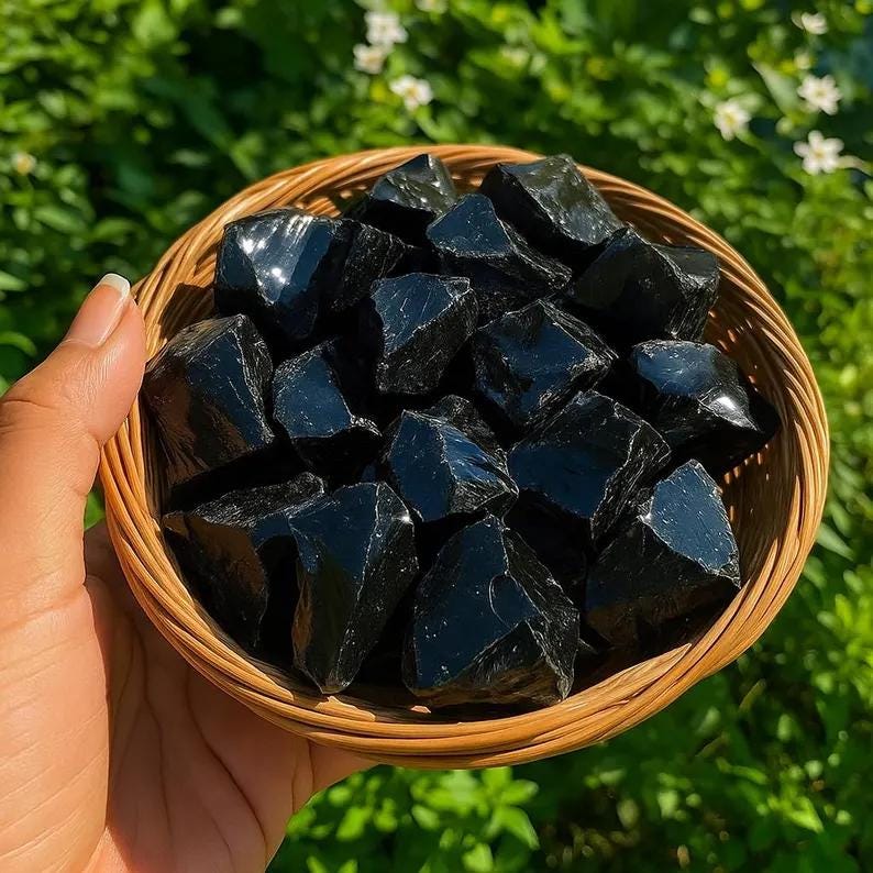 May include: A round, light brown woven wicker basket filled with numerous raw, glossy black obsidian stones. The stones have irregular, angular shapes with smooth, reflective surfaces that catch the light, creating sharp highlights across the dark texture.
