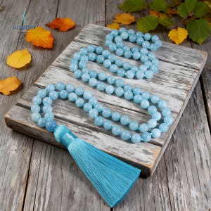 May include: Two aquamarine gemstone mala bead necklaces with a turquoise tassel rest on a weathered wooden surface. The necklaces are composed of round, light blue beads. The tassel is a vibrant turquoise color. Autumn leaves are scattered around the necklaces.
